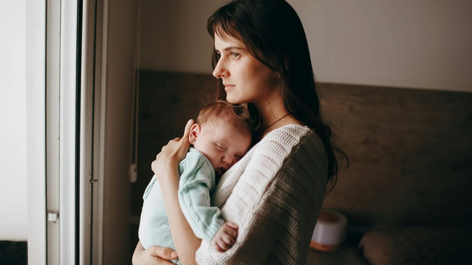 Mãe segura bebê recém-nascido adormecido no colo enquanto olha pela janela com expressão contemplativa em quarto com luz natural suave