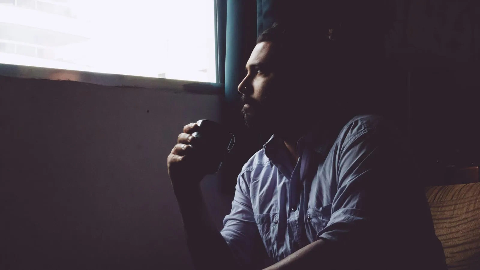 Homem de camisa clara segura caneca de café perto da janela, olhar contemplativo voltado pra luz natural do lado de fora