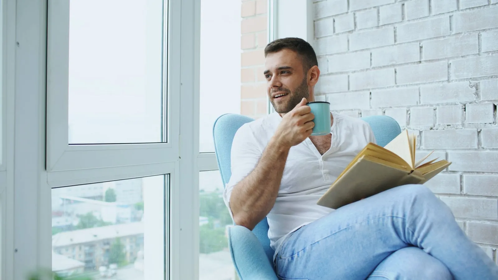 Homem relaxado lendo um livro com uma xícara de café ao lado, sentado perto de janela ampla com luz natural suave entrando no ambiente
