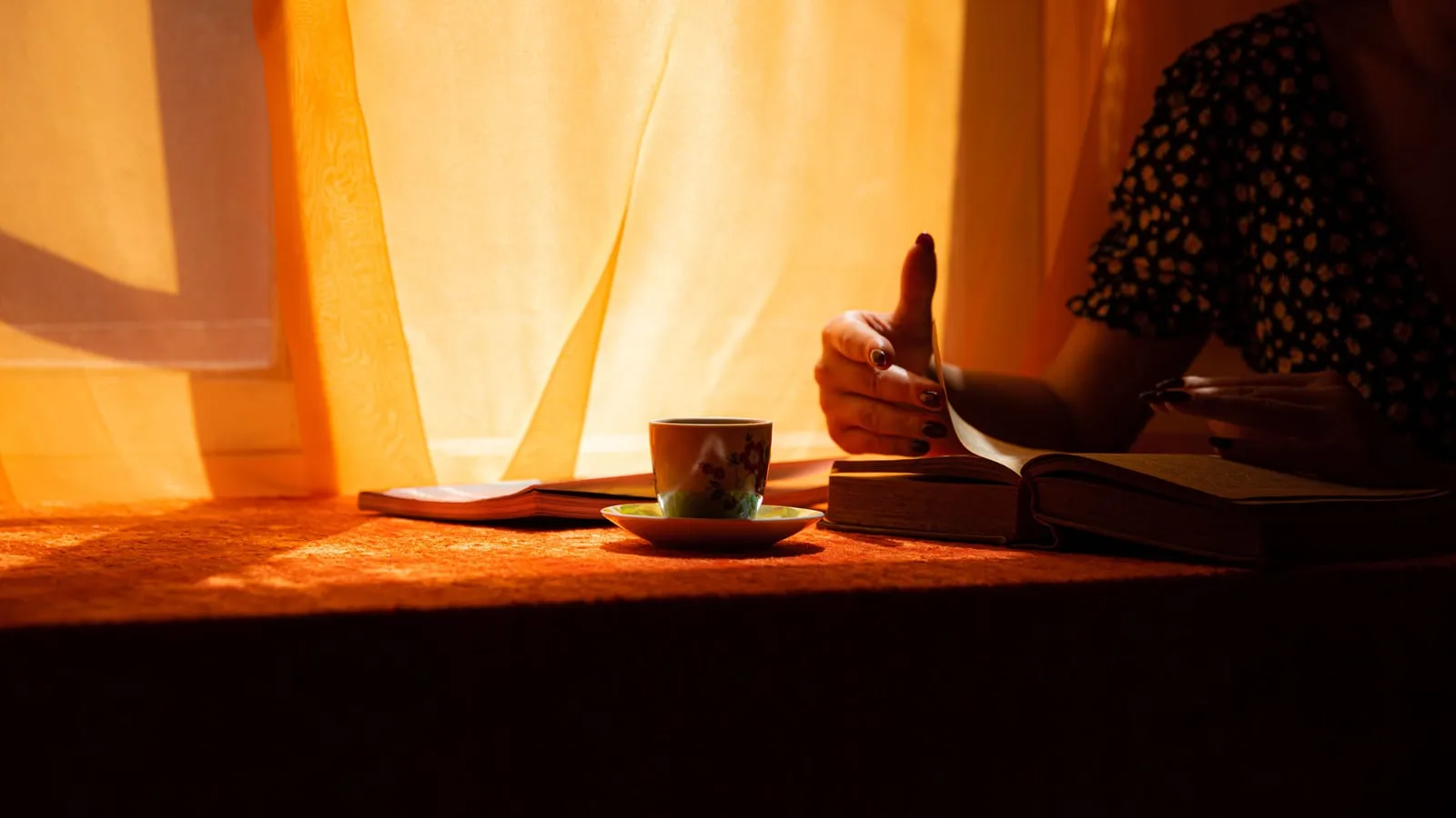 Pessoa lendo um livro segurando uma xícara de café perto da janela com luz natural suave