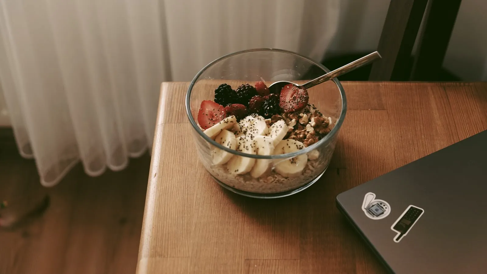 Tigela de vidro com aveia, banana, morango, amora, nozes e sementes de chia sobre mesa de madeira ao lado de um laptop, com luz natural de cortina ao fundo