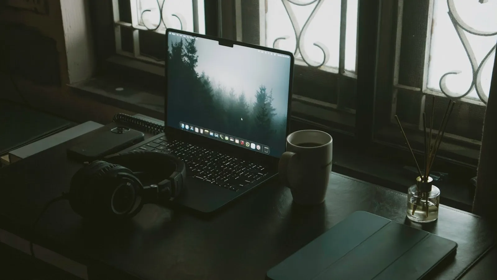 Setup de mesa doméstica com laptop, fones de ouvido e xícara de café junto à janela com luz natural