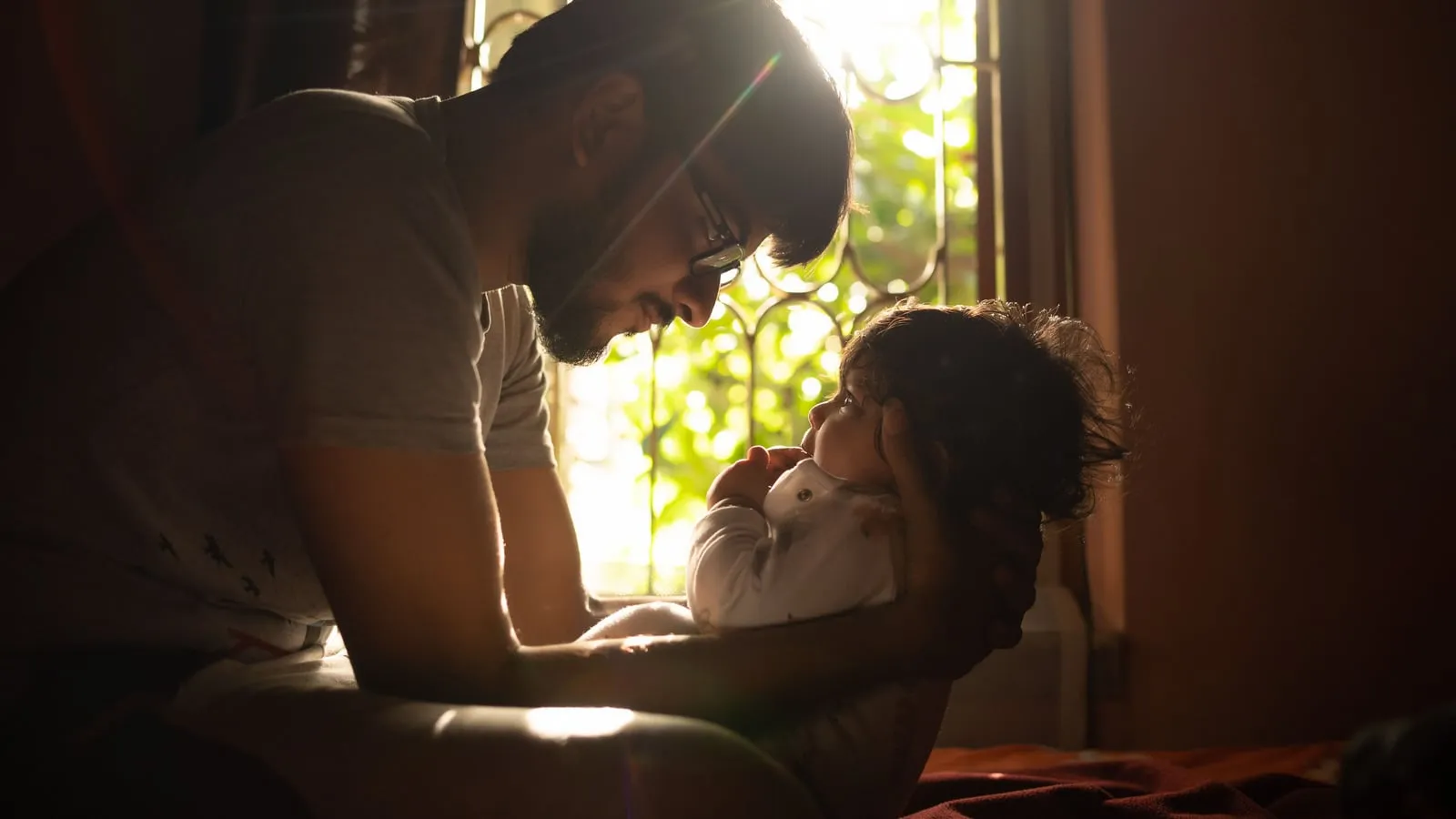 Pai segurando bebê no colo junto à janela, banhado por luz quente do final da tarde, em expressão contemplativa