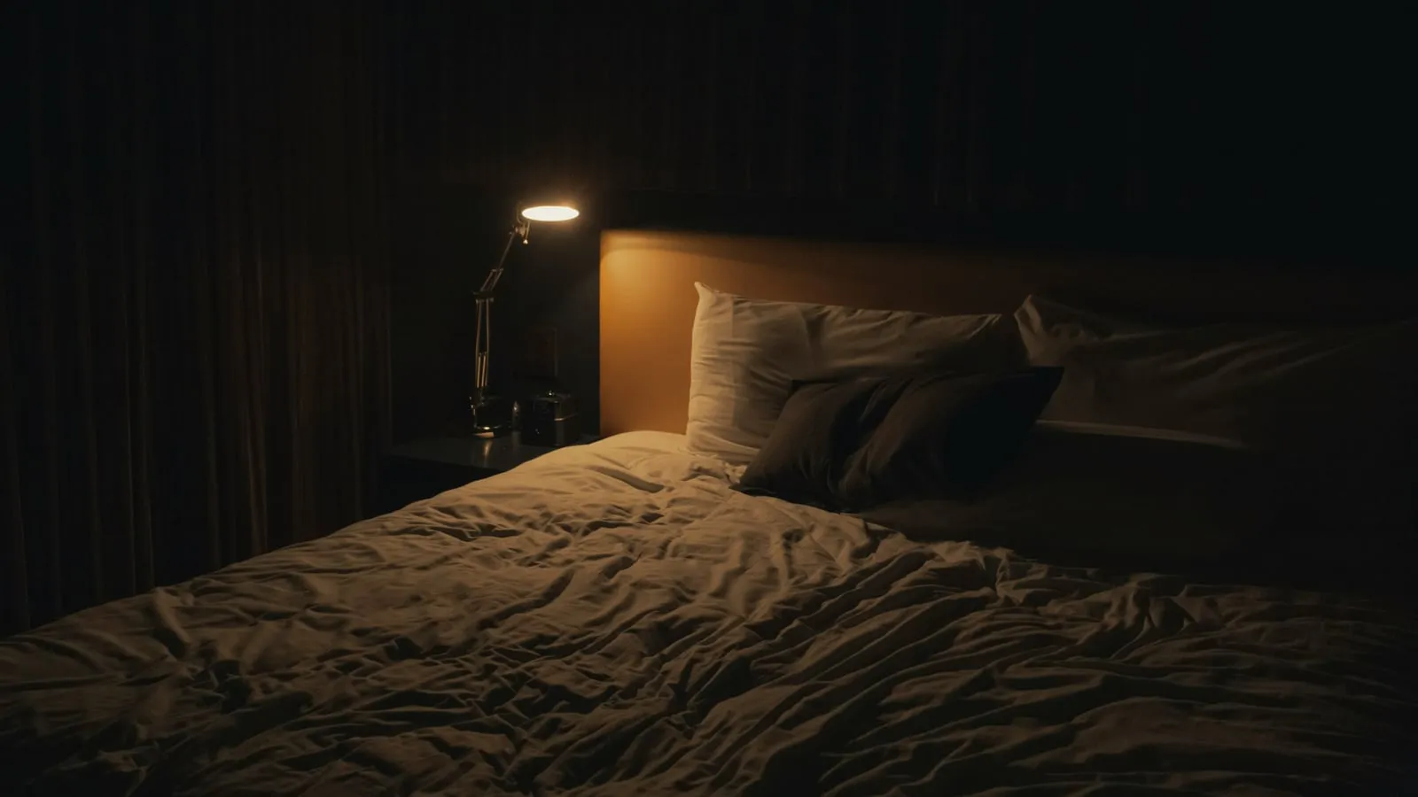 Abajur de cabeceira aceso com luz quente ao lado de cama arrumada, criando atmosfera serena de quarto noturno acolhedor