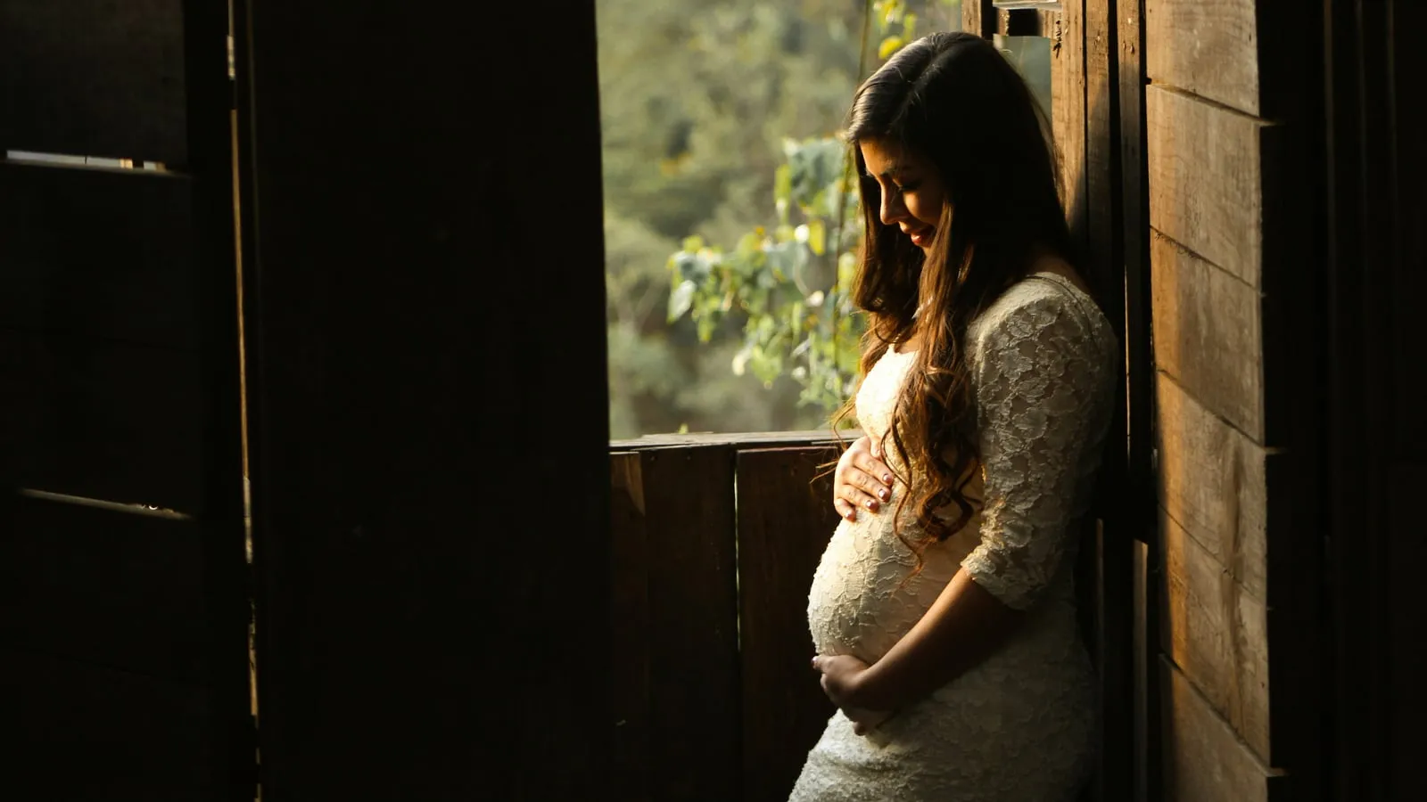 Mulher grávida com as mãos sobre a barriga, olhar voltado para baixo em atitude contemplativa, junto a uma janela de madeira com luz dourada natural