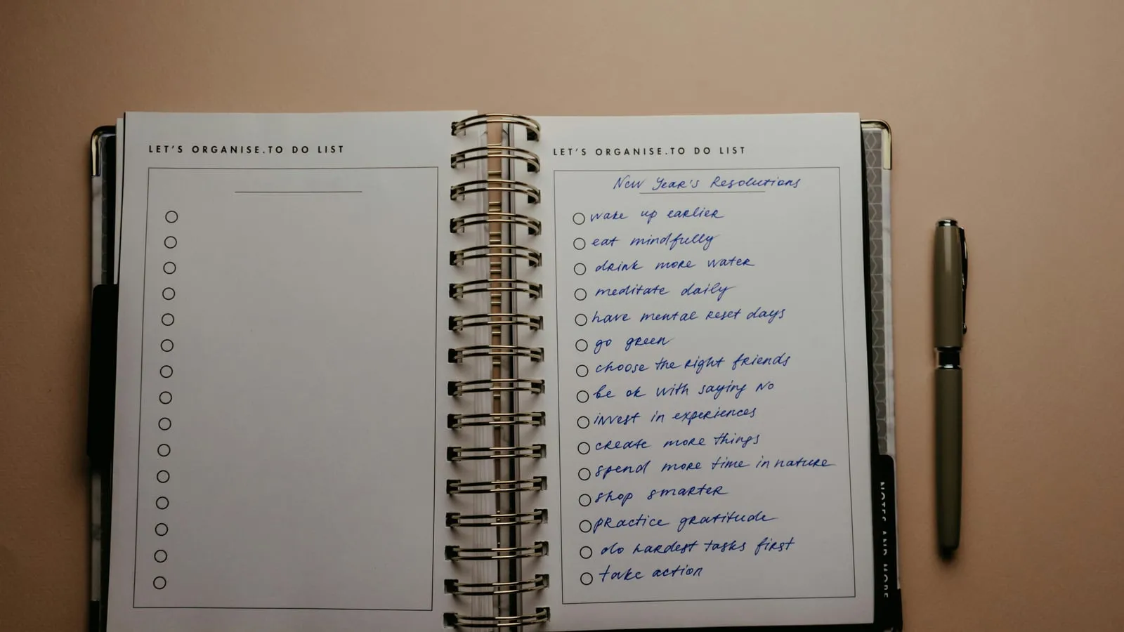 Planner espiral aberto sobre fundo bege, com lista de hábitos escritos à mão em tinta azul: 'wake up earlier', 'eat mindfully', 'meditate daily' entre outros. Uma caneta prata está apoiada à direita.