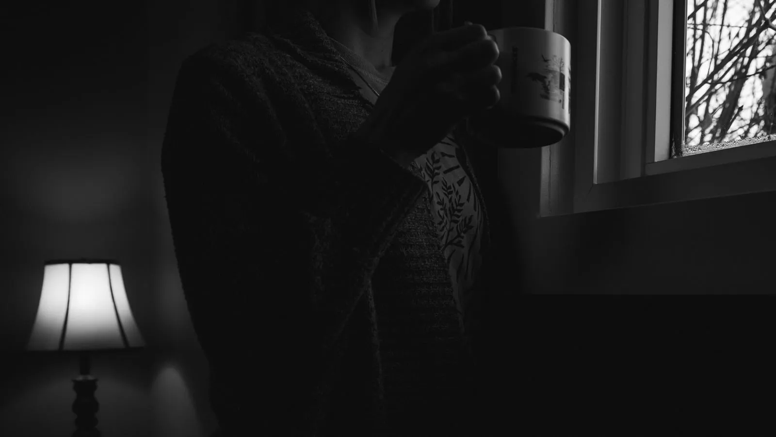 Mulher madura em postura contemplativa junto à janela, segurando xícara de café com luz quente da manhã iluminando o rosto