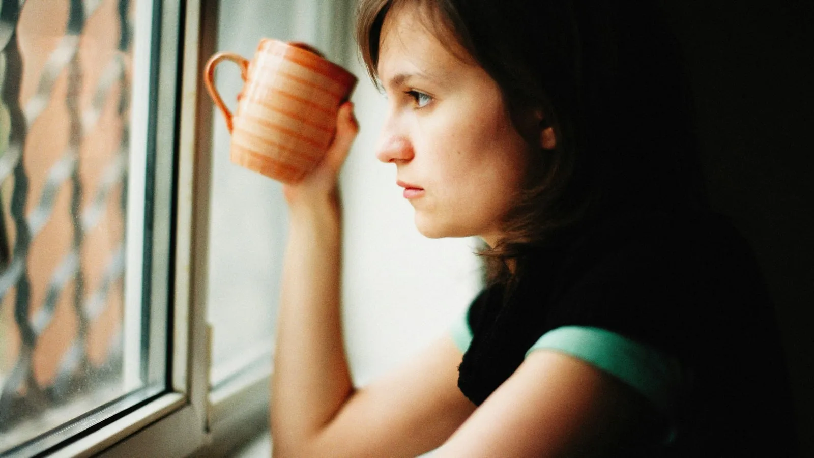 Mulher segurando xícara quente próxima à janela, em pausa contemplativa sob luz natural suave.