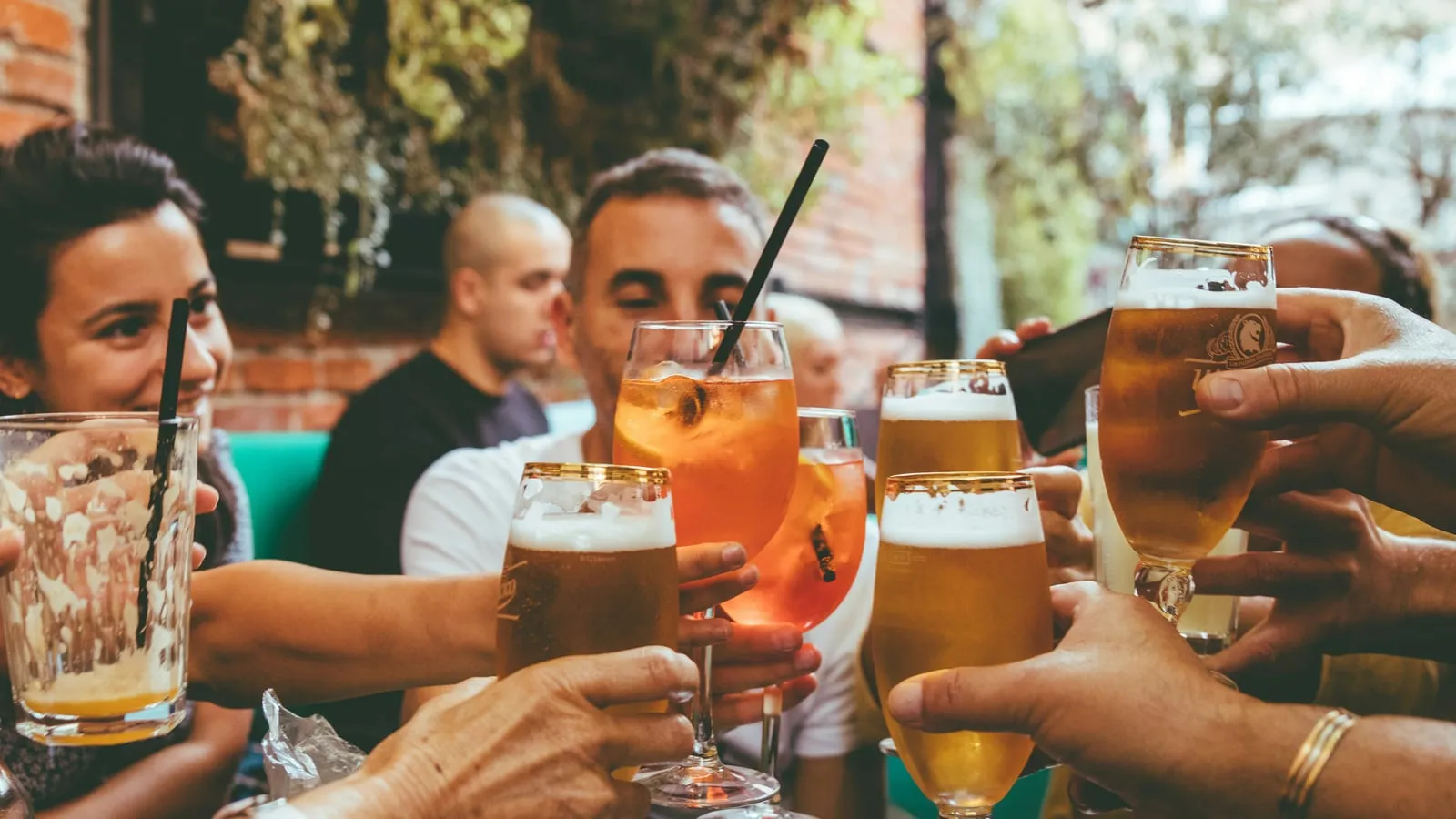 Grupo de amigos brinda com cervejas e drinks em mesa de barzinho ao ar livre, luz natural quente, clima social descontraído