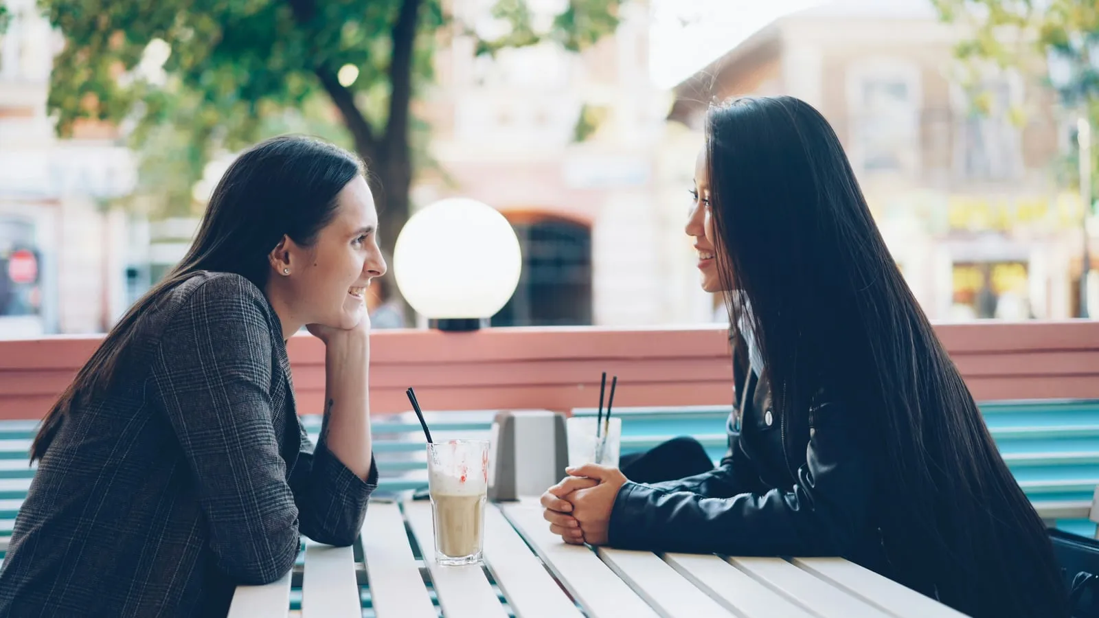 Duas mulheres conversam de frente uma para a outra em mesa de café ao ar livre, com contato visual atento e postura inclinada à frente