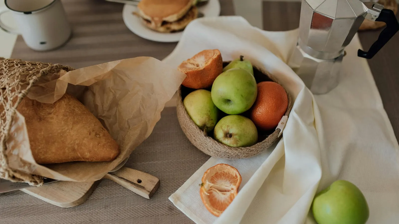 Mesa com cesta de frutas (maçãs verdes, pera, tangerina), pão rústico em papel kraft, cafeteira italiana e panquecas ao lado, em luz natural.