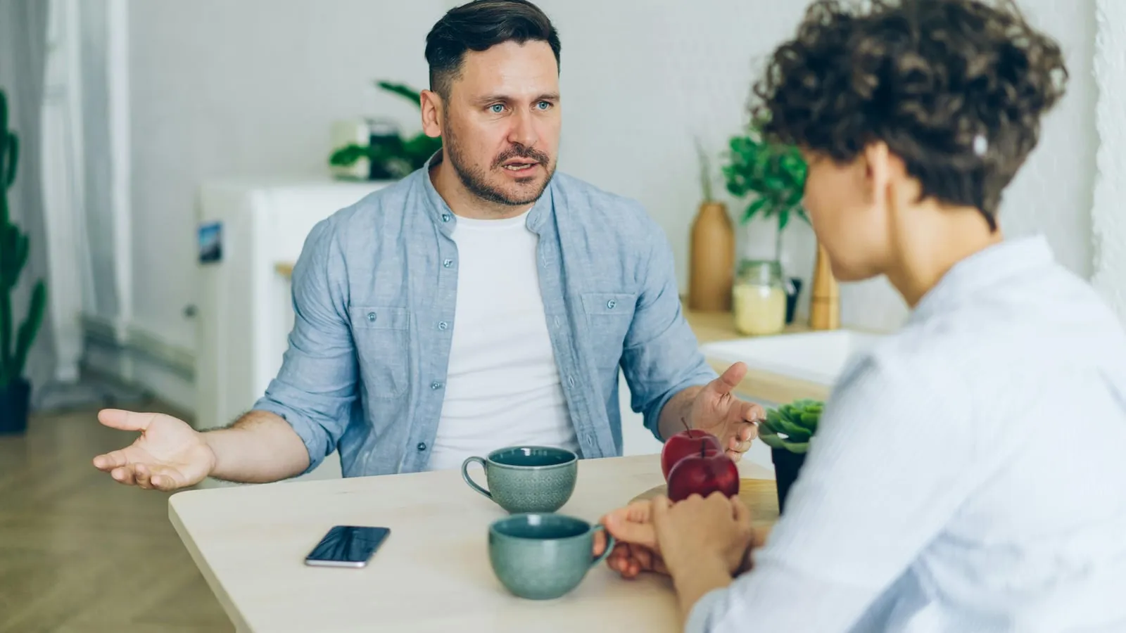 Duas pessoas adultas conversando em mesa de cozinha com xícaras de café entre elas, em luz natural quente.