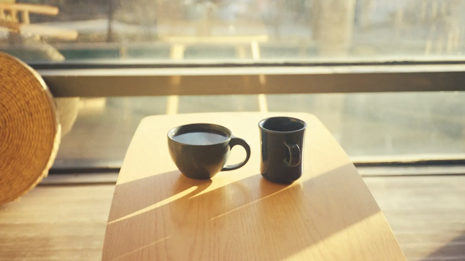 Duas xícaras de café lado a lado sobre mesa de madeira diante de janela com luz natural quente, sugerindo conversa pendente