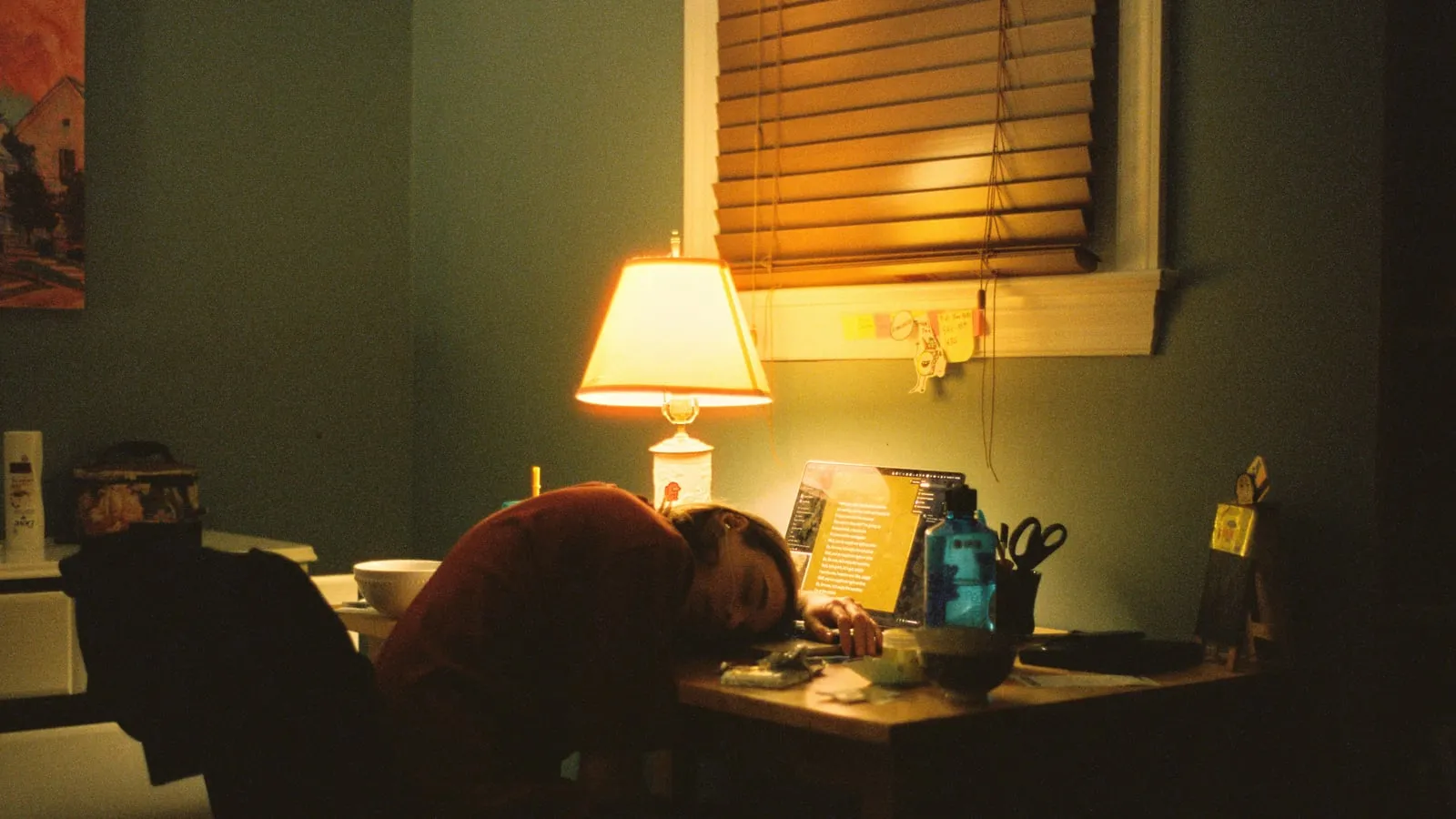 Pessoa com a cabeça apoiada na mesa ao lado do laptop, sob luz quente de abajur, em um quarto à noite.