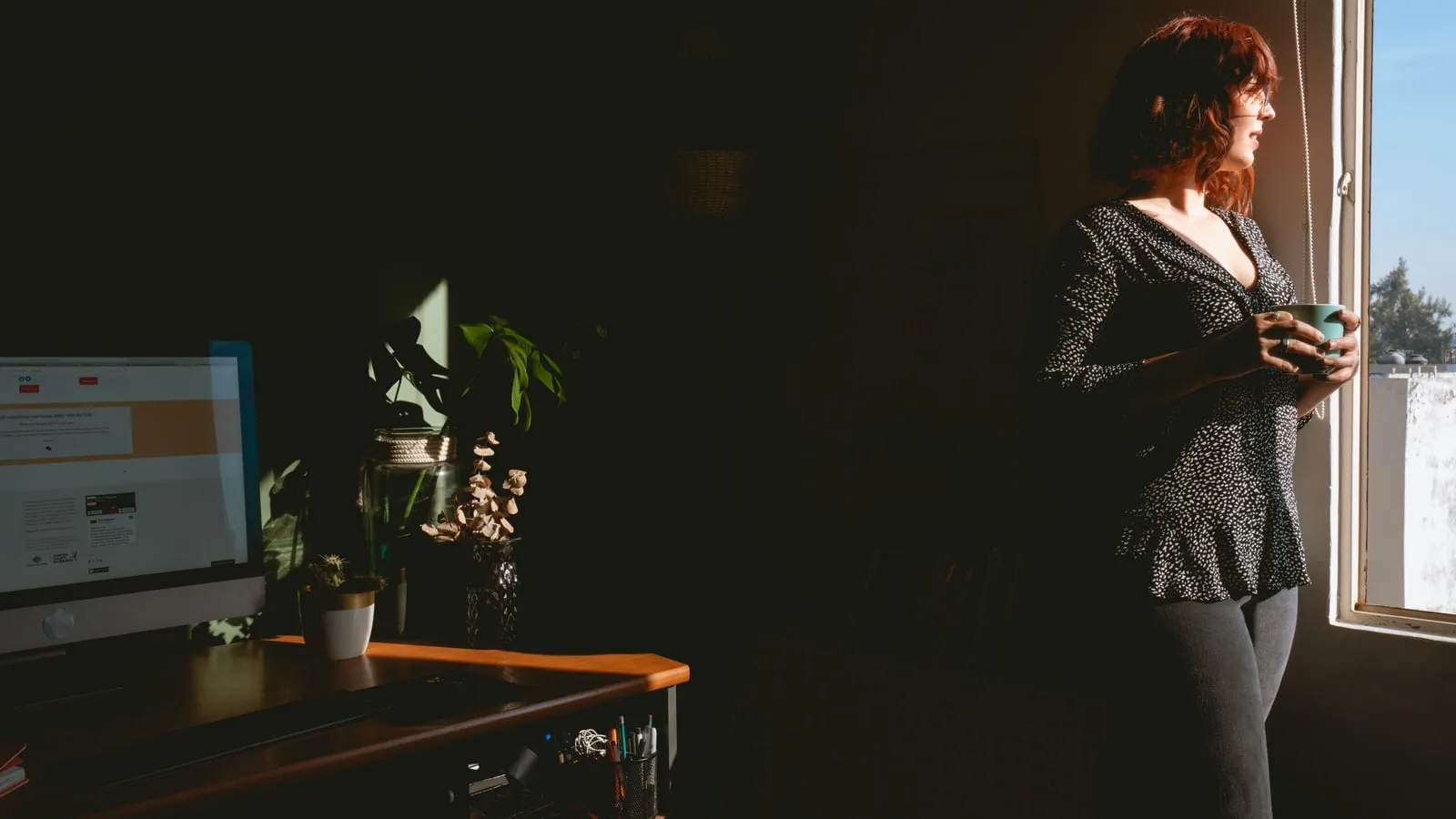 Mulher segurando xícara de café entre as mãos em casa, banhada por luz suave da manhã que entra pela janela