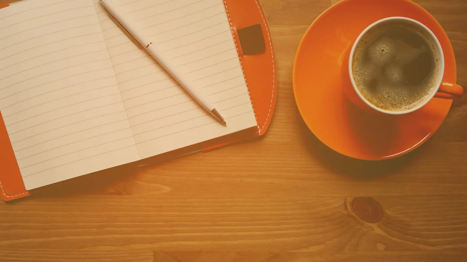 Caderno aberto com caneta apoiada sobre as folhas em branco ao lado de xícara de café em mesa de madeira clara, vista de cima