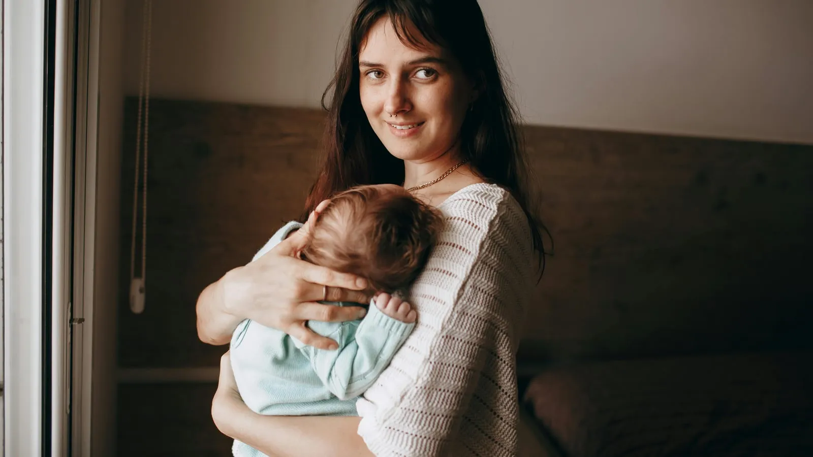 Mãe segurando bebê recém-nascido aninhado no ombro, em momento de pausa serena perto da janela com luz natural quente